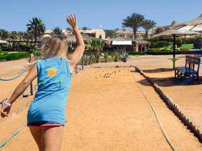 Osoba rzuca kulą do gry boccia na słonecznym piaszczystym boisku obok palm i parasoli.