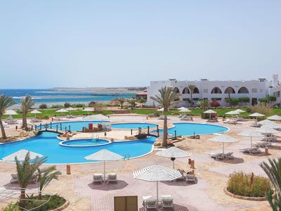 Large outdoor pool with sun umbrellas and loungers, white buildings and sea in the background.