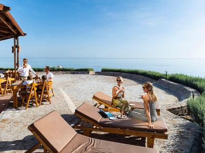 People relax on sun loungers and at a wooden table in an open, sunny beach bar.