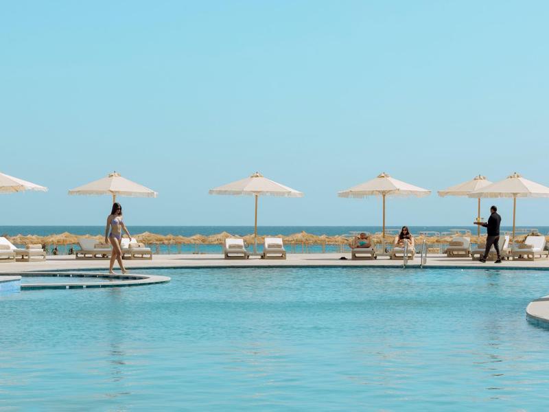 Clear blue pool with sun loungers and umbrellas under a bright sky beside the sea.