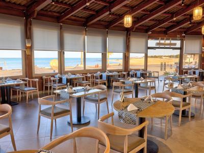 Ruime eetzaal met houten stoelen en tafels met uitzicht op het strand door grote ramen.