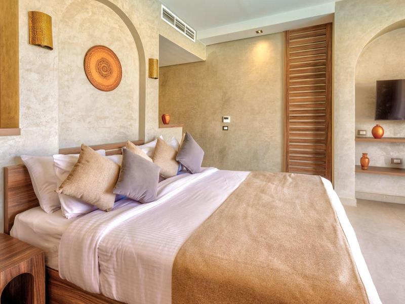 Helles Schlafzimmer mit großem Bett, beiger Bettdecke, mehreren Kissen und Wanddekor aus Holz.