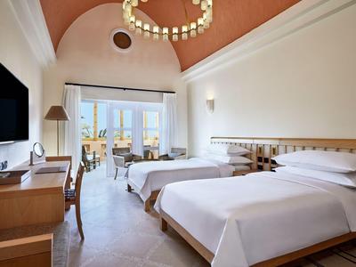 Helles Hotelzimmer mit zwei Einzelbetten, Schreibtisch, Fernseher und Balkon mit Meerblick.