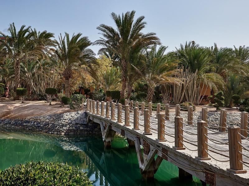 Holzbrücke mit Pfosten und Kordeln über türkisfarbene Lagune, Palmen und Sandstrand im Hintergrund.