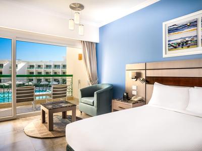 Helles Hotelzimmer mit Doppelbett, Balkon und Blick auf den Pool bei sonnigem Wetter.