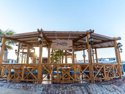 Offenes Holz-Pavillon am Hafen mit Blick auf Boote und Palmen bei klarem Himmel.
