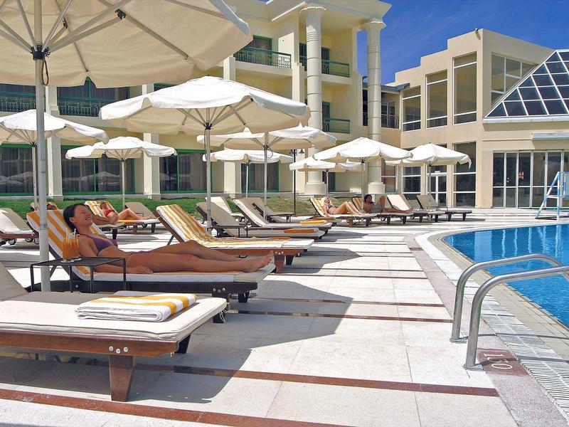 Poolbereich mit Liegestühlen, Sonnenschirmen und modernem Gebäude im Hintergrund an sonnigem Tag.