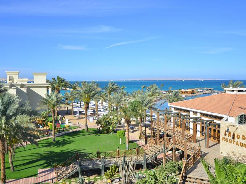 Blick auf Hotelanlage mit Palmen, grünen Rasenflächen und dahinter blaues Meer unter klarem Himmel.