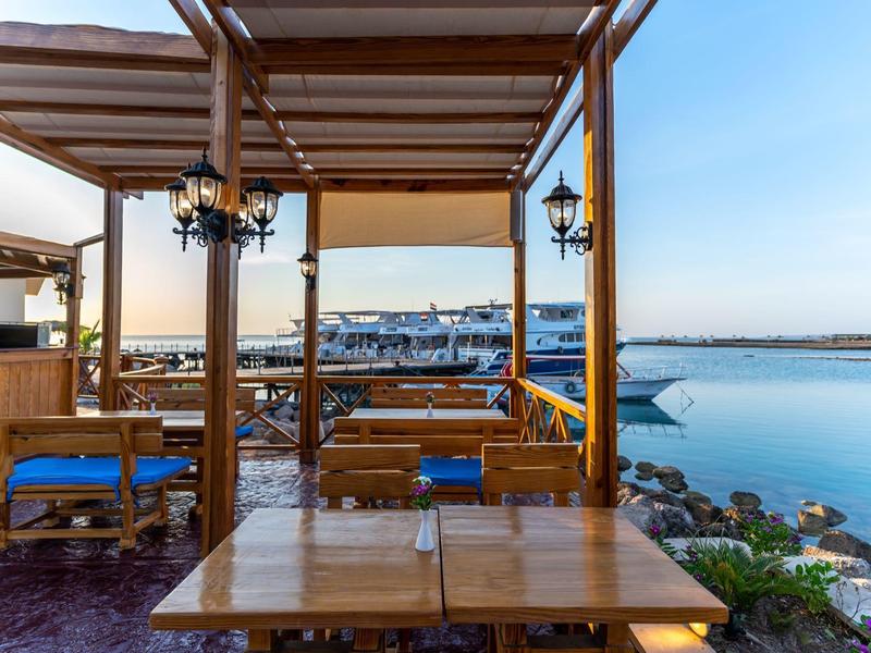 Blick von einer Terrasse mit Holztischen und blauen Stühlen auf einen Hafen und ruhiges Wasser.