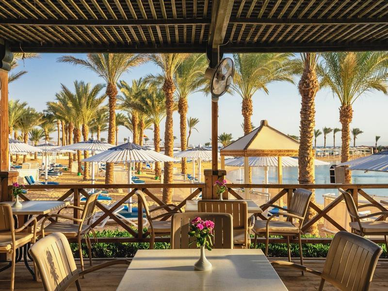 Terrassenblick mit Holzmöbeln, Palmen und Booten bei klarem Himmel am Wasser.