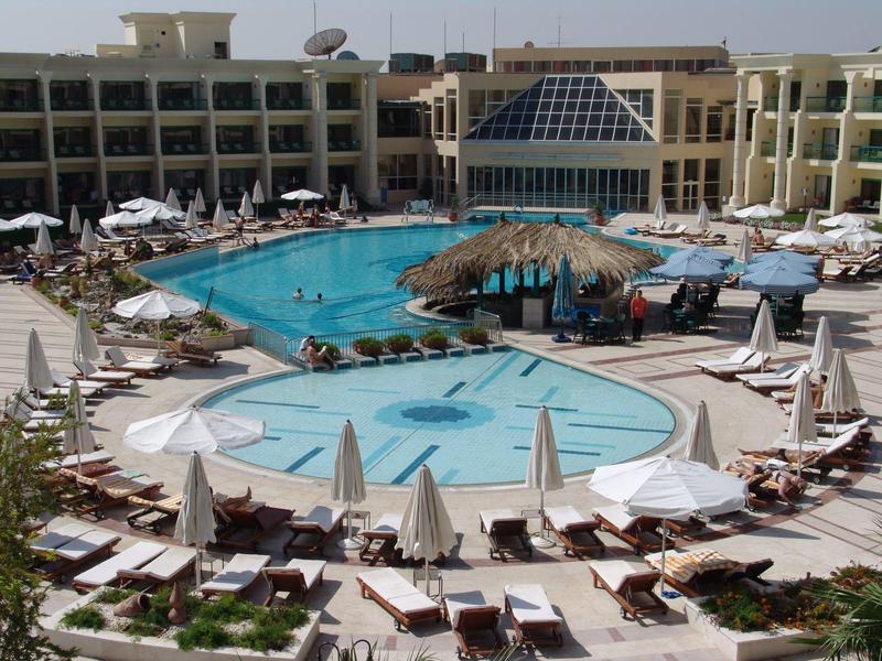 Großer Hotelpool mit runden Sonnenliegen, weißen Schirmen und mehrstöckigem Gebäude im Hintergrund.