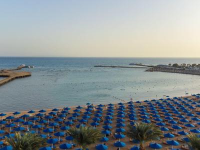 Strand mit vielen blauen Sonnenschirmen, ruhiges Meer und klarer Himmel im Hintergrund.