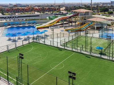 Freizeitzentrum mit grünem Fußballfeld, mehreren Schwimmbecken und Wasserrutschen bei sonnigem Wetter.