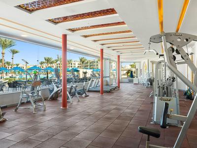 Großer, heller Fitnessraum mit Cardio- und Kraftgeräten, draußen Pool und Palmen sichtbar.