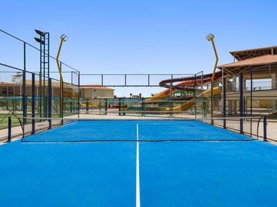 Blauer Padel-Tennisplatz im Freien mit Zaun und Gebäuden im Hintergrund unter klarem Himmel.