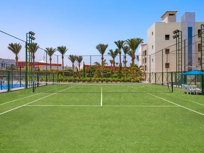 Grüner Tennisplatz mit Palmen, Zaun und Gebäuden im Hintergrund bei klarem Himmel.