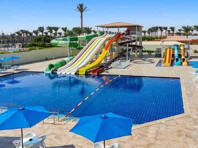 Wasserpark mit mehreren Wasserrutschen und einem großen Schwimmbecken, umgeben von Liegestühlen und blauen Sonnenschirmen.