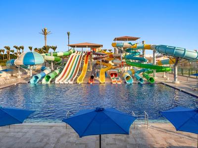 Wasserpark mit mehreren bunten Wasserrutschen, Pool und blauen Sonnenschirmen im Vordergrund.