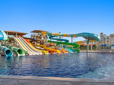Mehrere Wasserrutschen verschiedener Größen und Farben münden in einem Hotel-Pool unter klarem Himmel.