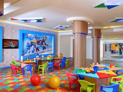 Buntes Kinderzimmer mit kleinen Tischen, Stühlen, Spielzeug und einer großen Wand mit Comicfiguren.