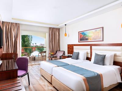 Helles Hotelzimmer mit zwei Betten, Holzmöbeln, Balkon und Blick auf Garten und Meer.