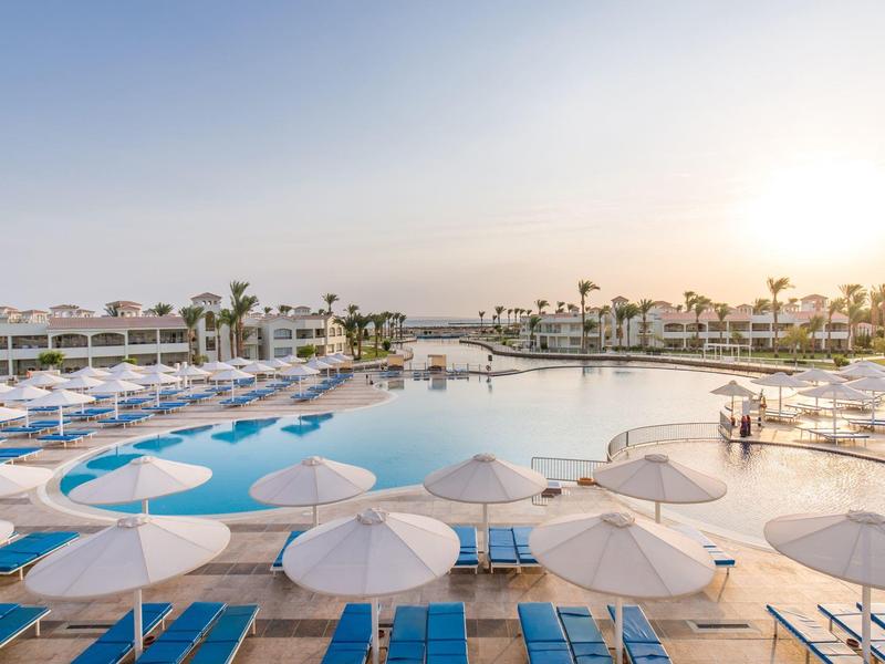 Großer Hotelpool mit weißen Sonnenschirmen und blauen Liegen bei Sonnenuntergang.