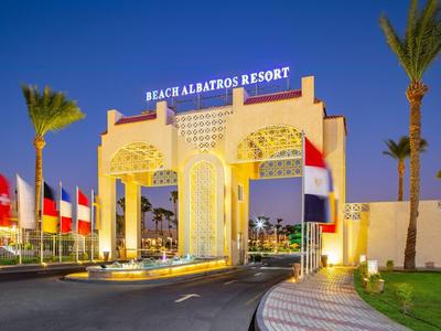 Eingangstor des Beach Albatros Resorts mit Beleuchtung, Palmen und Flaggen bei Abenddämmerung.