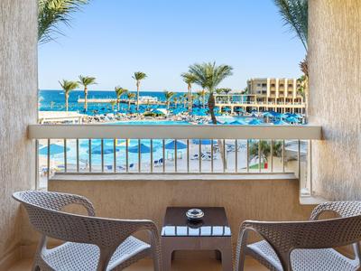 Balkon mit zwei Stühlen, Tisch und Blick auf Pool, Palmen und das Meer bei klarem Himmel.