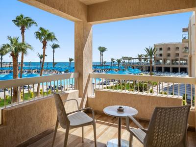 Balkon mit Tisch, zwei Stühlen und Blick auf Pool, Palmen und Hotel am Meer bei sonnigem Wetter.