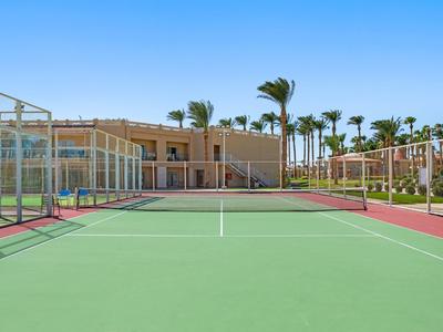 Tennisplatz mit grünem Belag, roten Außenlinien, Palmen und Gebäuden im Hintergrund bei klarem Himmel.
