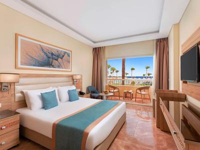 Helles Hotelzimmer mit Doppelbett, blauen Akzentkissen, Balkon und Meerblick, warme Holzmöbel.