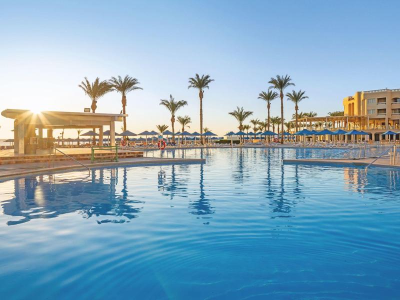 Schwimmbad bei Sonnenuntergang mit Palmen und Hotelgebäude im Hintergrund.