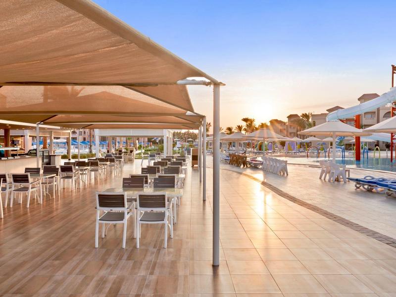 Moderner Außenbereich mit Holzboden, weißen Tischen, Stühlen und Sonnenschirmen bei Sonnenuntergang.