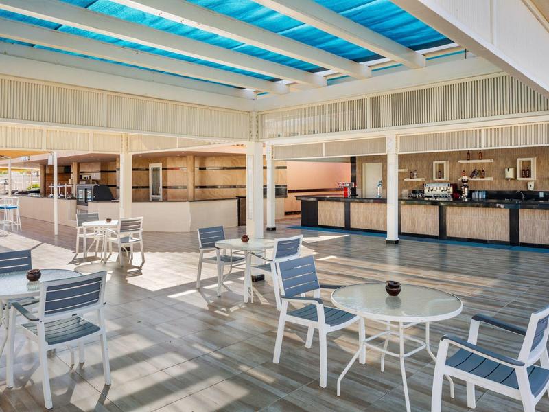 Helle, offene Terrasse mit weißen Tischen und Stühlen unter einem glasüberdachten Dach.