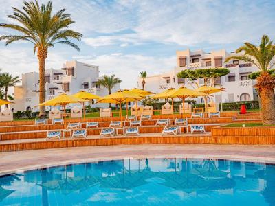 Rundlicher Pool vor Reihen mit Sonnenschirmen und Liegestühlen bei Hotelgebäuden und Palmen.
