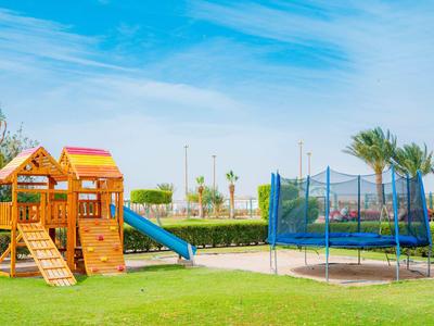 Kinderspielplatz mit Holzhaus, Rutsche und Trampolin auf grünem Rasen unter blauem Himmel.