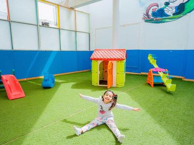 Kind sitzt auf grünem Kunstrasen in einem bunten Indoor-Spielplatz mit Rutsche, Haus und Schaukeln.