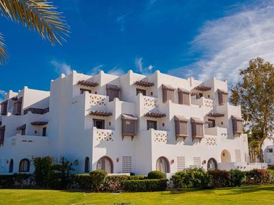 Weißes Hotelgebäude mit Balkonen unter blauem Himmel, umgeben von grüner Wiese und Bäumen