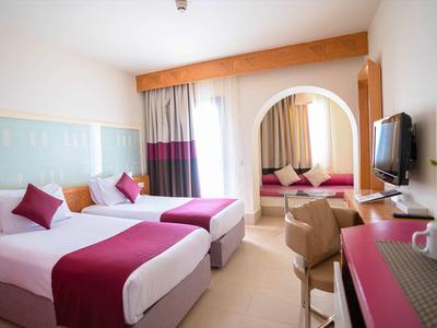 Helles Hotelzimmer mit zwei Betten, lila Bettwäsche, Spiegel, Fernseher und einem Schreibtisch.