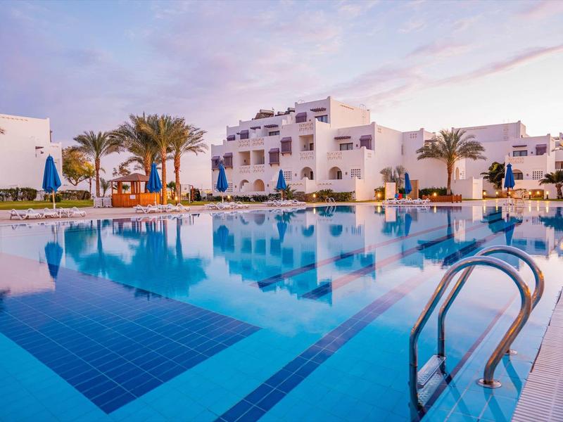 Moderner Hotelkomplex mit weißem Gebäude und großem blauem Pool bei Sonnenuntergang.