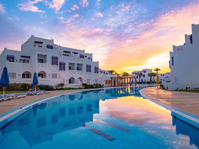 Ein blauer Pool zwischen weißen Gebäuden und buntem Himmel bei Sonnenuntergang.