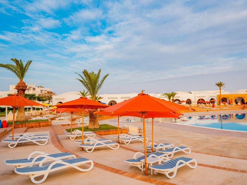 Poolbereich mit weißen Liegestühlen und orangen Sonnenschirmen unter blauem Himmel.