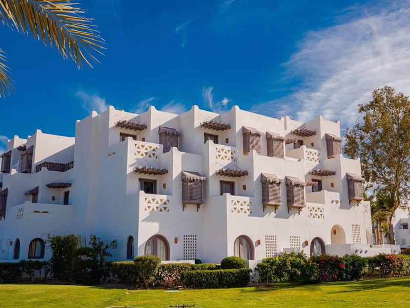 Weißes mehrstöckiges Gebäude mit Balkonen, blauer Himmel, Palmen und grüner Rasenfläche im Vordergrund.
