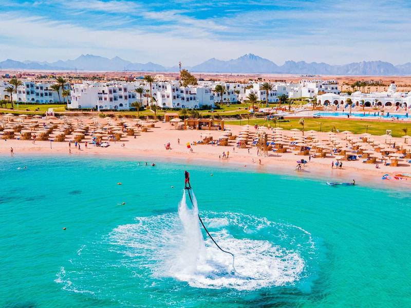 Person fliegt mit Wasserdruckgerät über türkisfarbenem Meer vor Sandstrand und Hotelanlage.