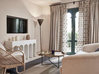 Helles Wohnzimmer mit weißen Möbeln, großem Fenster, Vorhängen und modernem Holzsessel.