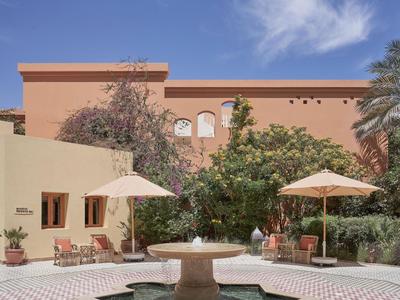 Innenhof mit Brunnen, Palmen und Sonnenschirmen vor einem rosafarbenen Gebäude unter blauem Himmel.