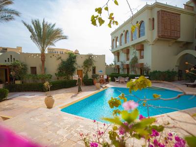 Ein sonniger Innenhof mit blauem Pool, Palmen, Pflanzen und einem zweistöckigen Gebäude.