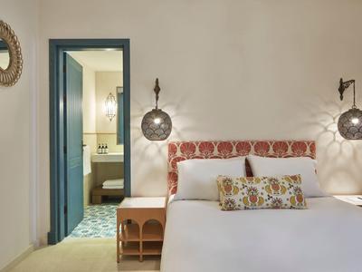 Schlafzimmer mit weißem Bett, gemustertem Kopfteil, Nachttischen und blauem Türrahmen zum Bad.