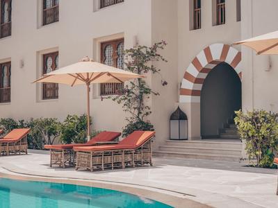 Poolbereich mit Liegestühlen und Sonnenschirmen vor einem weißen Gebäude mit Bogen