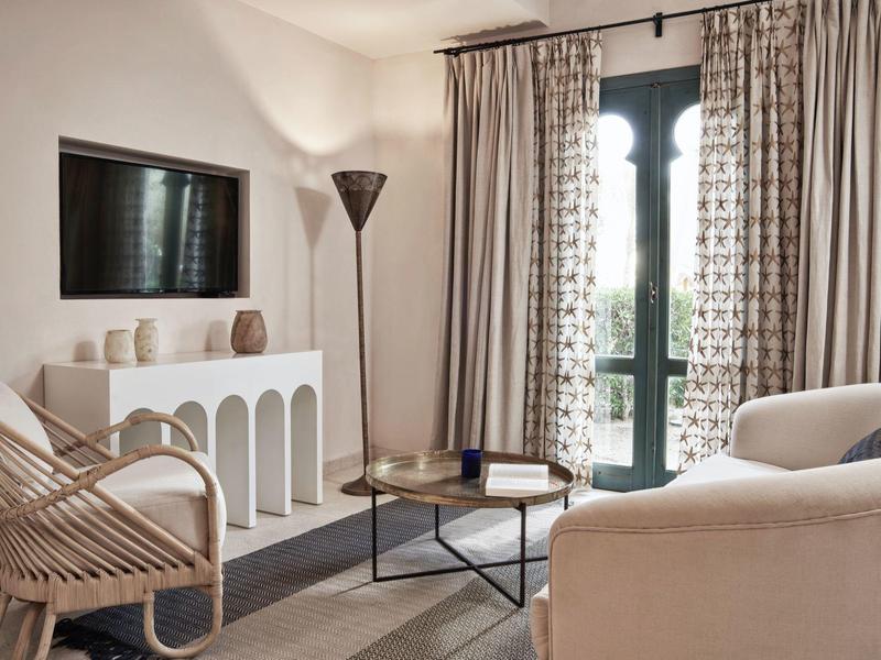 Helles Wohnzimmer mit weißen Möbeln, großem Fenster, Vorhängen und modernem Holzsessel.